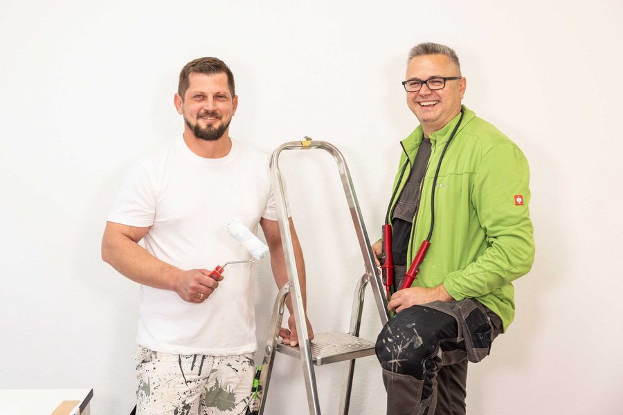  Zwei Männer, in Arbeitskleidung, einer in grüner Jacke, stehen nebeneinander mit einer Leiter in der Mitte.
